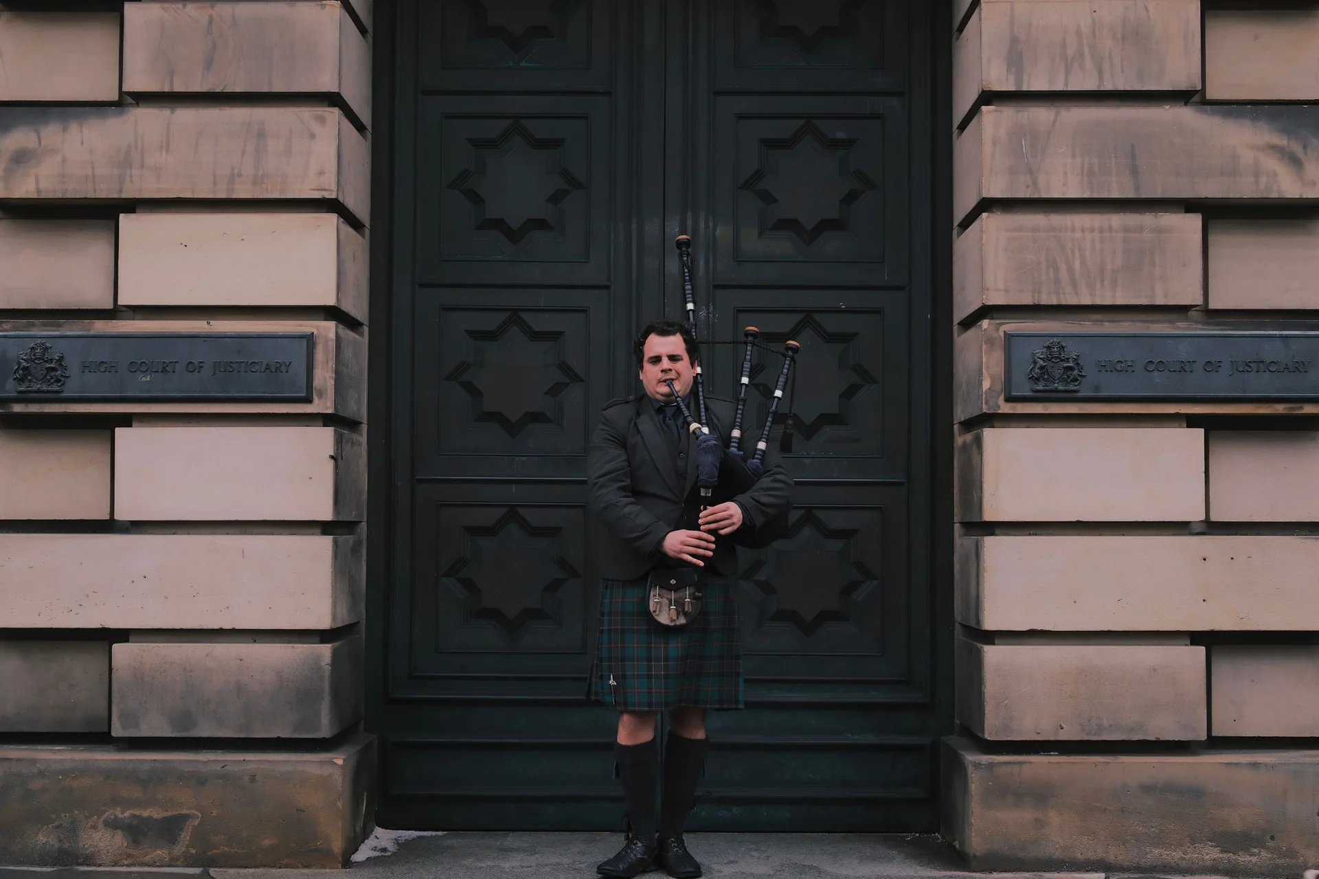 A piper in full Highland dress playing bagpipes outside a grand stone doorway in Edinburgh